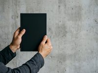Person of South Asian descent holding blank black book against textured concrete wall, wearing dark long-sleeved shirt, suitable for publishing, education, and branding mockups.