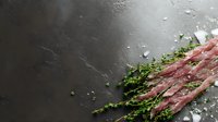 Raw prosciutto slices with fresh thyme sprigs and sea salt crystals arranged on dark slate stone background, dramatic side lighting creates texture.