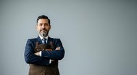 Mature male professional with dark hair and beard wearing navy blazer, light blue shirt, and brown vest, arms crossed in confident pose against neutral gray background.