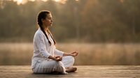 Peaceful young woman meditating in lotus position on wooden dock at sunrise, practicing mindfulness and yoga outdoors in serene natural setting.