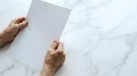 Senior adult hands holding blank white paper on marble surface for mockup and design presentations.
