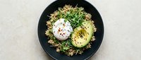 Overhead view of a black plate with quinoa salad, sliced avocado, poached egg, and fresh herbs on light background. Perfect for food blogs and healthy eating content.