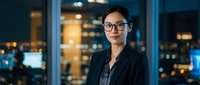 Professional Asian businesswoman in glasses and dark suit standing confidently in modern office with city lights background at night for corporate presentations.