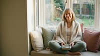 Mature blonde woman in cozy cardigan relaxing on sofa by window with book, peaceful home interior lifestyle for wellness and leisure reading concepts.