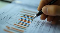 Close-up of hand with pen analyzing colorful bar graph data on financial report, showing business performance metrics for investment decision-making.