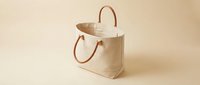 Neutral canvas tote bag with brown leather handles on beige background, suitable for retail, fashion, and e-commerce product photography.