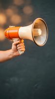 Hand holding orange and white megaphone bullhorn against blurred bokeh background for communication marketing and announcement campaigns.