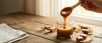 Hand drizzling golden caramel sauce from wooden spoon into glass jar on rustic wooden table with scattered caramel candies in warm natural lighting.