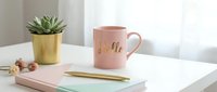 Pink ceramic mug with gold hello lettering on white desk with succulent plant, notebook and gold pen, cozy feminine workspace.