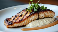 Grilled salmon fillet with char marks served on creamy cauliflower puree, garnished with fresh microgreens and sauce, presented on white plate against dark background.