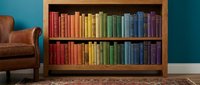 Wooden bookshelf filled with rainbow-colored books arranged in gradient order, set against a teal wall with a leather armchair beside it.