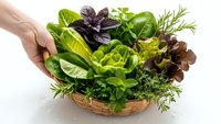 Fresh mixed herbs and leafy greens in wicker basket held by hand on white background. Organic cooking ingredients for healthy meals and recipes.