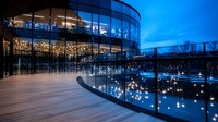 Modern glass building illuminated at dusk with curved architecture reflecting blue twilight sky, wooden deck foreground.