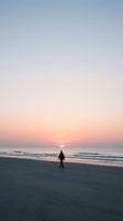Solitary figure walking on empty beach during golden hour sunset with pastel sky colors and gentle ocean waves for peaceful meditation and wellness concepts.