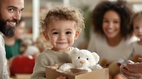 Happy toddler with curly hair holding white teddy bear, smiling at camera while sitting with diverse family at home during cozy gathering.