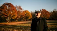 Mature man with beard wearing cap and dark jacket standing in autumn field with golden trees and clear sky, suitable for lifestyle, seasonal, and outdoor editorial use.