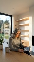 Mature woman with glasses working on tablet at modern home office desk with floating shelves and natural light from large window.