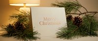 Merry Christmas greeting card with gold foil lettering surrounded by pine branches and cones in warm glowing light, festive holiday decor.