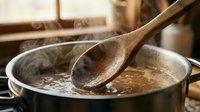 Wooden spoon stirring hot broth or soup in metal pot with steam rising, suitable for cooking, food preparation, and culinary content.