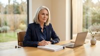 Professional mature blonde businesswoman in dark suit writing notes at modern office desk with laptop and natural lighting from large windows.