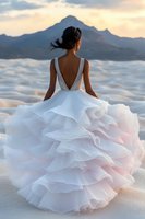 Young African American bride in luxurious white ruffled wedding dress with deep V-back stands against mountain backdrop during sunset, her hair flowing in wind.