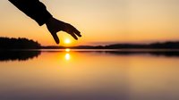 Hand reaching toward glowing sunset over calm lake, silhouette against golden sky reflected on water, nature and hope concept.