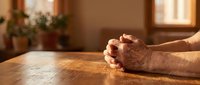 Elderly hands clasped in prayer on wooden table in warm sunlit room with soft lighting creating peaceful spiritual atmosphere for meditation and faith.