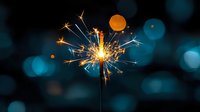 Glowing sparkler emitting bright sparks against dark background with blue bokeh lights creates festive holiday atmosphere. Celebration moment captured in detail.