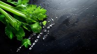 Fresh green celery stalks and leaves with scattered sea salt crystals on wet black stone background, dramatic lighting creates contrast for food photography.