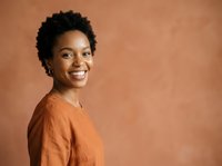 Young African American woman with short natural hair wearing an orange shirt, smiling at camera against warm terracotta background for portrait and lifestyle use.