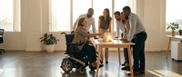 Diverse business team including wheelchair user collaborating around table in bright modern office space promoting workplace inclusion and accessibility.