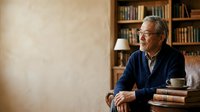 Senior man with gray hair and glasses, East Asian ethnicity, wearing navy blue cardigan, sitting thoughtfully in home library with bookshelves, coffee cup and stacked books nearby for education and lifestyle content.