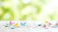 Colorful paper butterflies scattered on white surface with soft green bokeh background for spring crafts and decoration projects.
