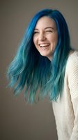 Young woman with vibrant blue-green dyed hair laughing joyfully in white sweater against neutral background for lifestyle and fashion content.