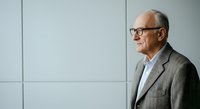 Senior man with gray hair and glasses wearing a gray blazer and white shirt, standing in profile against a white paneled wall in a modern office or corporate setting.