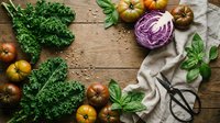 Fresh organic vegetables including kale, purple cabbage, heirloom tomatoes and basil herbs arranged on rustic wooden table for healthy cooking and meal preparation.