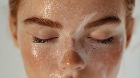 Young woman with freckles and natural glowing skin in closeup portrait with eyes closed, showing dewy complexion and natural beauty in soft lighting.