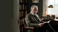 Elderly Caucasian man with gray beard reading book in cozy home library with cat and lamp, evoking wisdom, solitude, and intellectual lifestyle.