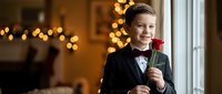 Young boy in elegant black tuxedo holding red rose by window with warm bokeh lights creating romantic atmosphere for special occasions and formal events.