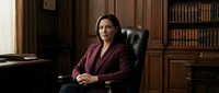 Professional businesswoman in burgundy blazer sitting confidently in executive leather chair in elegant wood-paneled office with bookshelf background.