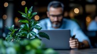 Young professional working on laptop in cozy evening atmosphere with green plant in foreground, holding coffee cup, bokeh lights background creates warm ambiance.