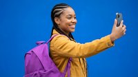 Young African American woman with braids taking selfie with smartphone wearing yellow jacket and purple backpack on blue background.