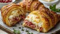 Fresh baked ham and cheese croissant sandwich cut in half showing melted cheese and meat filling, garnished with herbs on rustic wooden board.