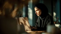 Young Middle Eastern woman in hijab working on laptop computer in modern office with warm natural lighting and professional atmosphere.