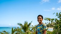 Young South Asian male wearing sunglasses and patterned shirt standing outdoors with tropical beach, palm trees and ocean in background on sunny day.