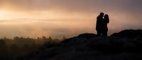 Romantic couple silhouette embracing on mountain peak during golden sunset with misty valley landscape below for love and adventure concepts.