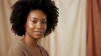 Young African American woman with natural curly afro hair smiling warmly at camera in cozy indoor setting with soft lighting and neutral tones.