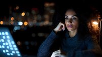 Thoughtful young African American woman with curly hair working late on computer with city lights in background, contemplative expression.