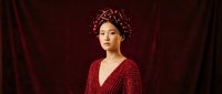 Woman with East Asian appearance wearing deep red polka dot dress and ornate floral crown against burgundy velvet backdrop for portrait and editorial use.