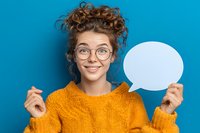 Young woman with curly hair and glasses holding blank speech bubble against blue background, perfect for communication concepts.